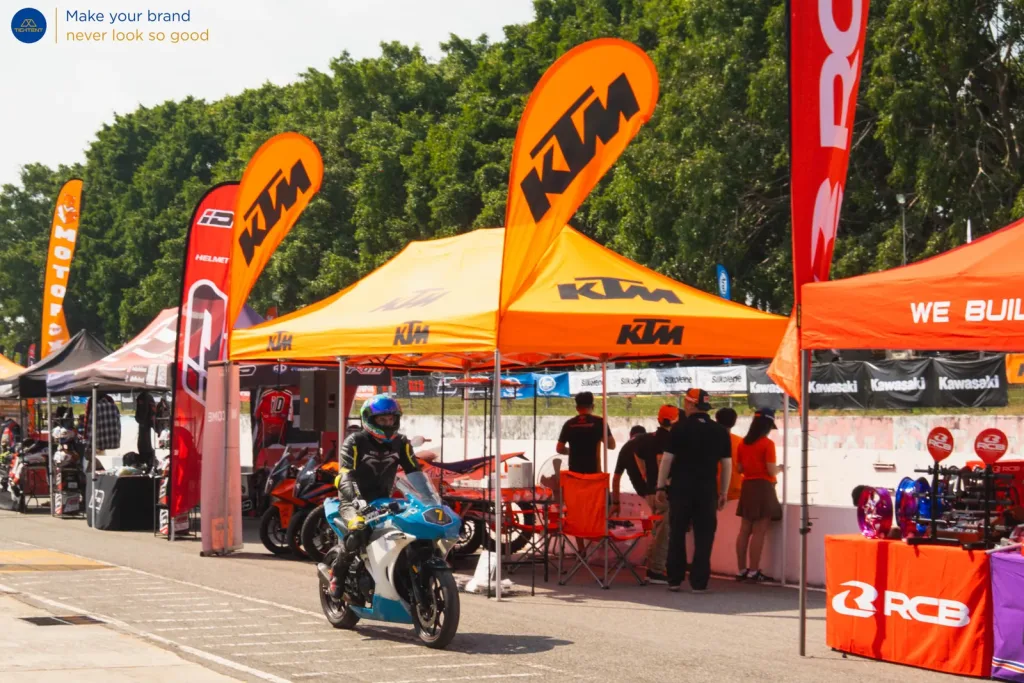 เต็นท์พับสีส้ม KTM และธงปีกนก ผู้ขับขี่ผ่านหน้าบูธ